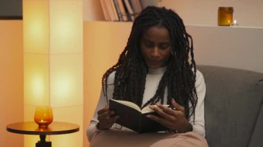 Akşamları kanepede kitap okuyan genç Afrikalı Amerikalı kadın sıcak ve rahat bir ev atmosferinde parlayan bir mumun yanında sakin bir odak noktasıyla sayfaları çeviriyor. Yüksek kalite 4k görüntü