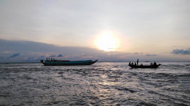 Suyun ya da nehrin üzerinde huzurlu bir günbatımı. Biri daha büyük, biri daha küçük iki tekne görülebilir. Parçalı bulutlu gökyüzü ve sıcak parıltı sakin bir atmosfer yaratır..