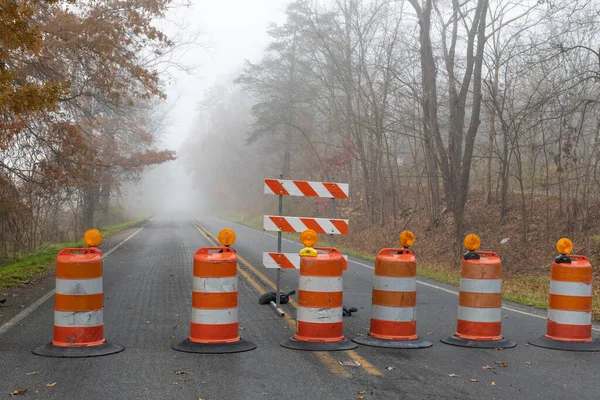 Row of tightly spaced orange traffic barrels barricading a road disappearing into a foggy autum landscape, horizontal aspect