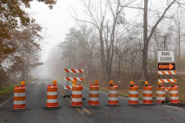 Straight road disappearing into a dense fog, barricade of bright orange traffic barrels, horizontal aspect