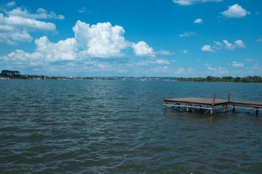 Brezilya, Braslia 'da Parano Gölü manzaralı, ön planda ahşap bir rıhtım, durgun sular, berrak mavi bir gökyüzü, ve uzaktan görünen ikonik JK Köprüsü.