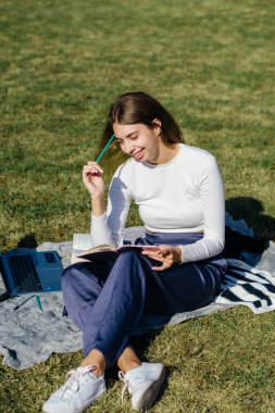 student girl is studying outside at the summer Holliday at the university on the green grass using laptop and taking notes clipart