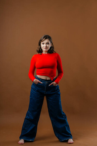 A fashionable young woman in a bold red top and wide-leg denim jeans strikes a dynamic and artistic pose against a warm brown background. High quality photo