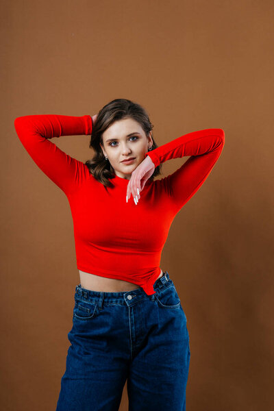 A fashionable young woman in a bold red top and wide-leg denim jeans strikes a dynamic and artistic pose against a warm brown background. High quality photo