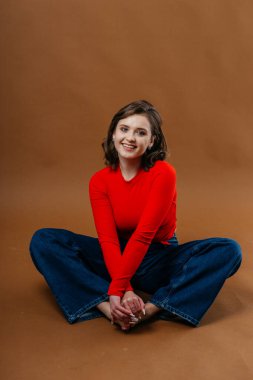 A fashionable young woman in a bold red top and wide-leg denim jeans strikes a dynamic and artistic pose against a warm brown background. High quality photo clipart