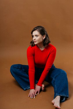 Kalın kırmızı bluzlu ve geniş paçalı kot pantolonlu şık bir genç kadın, dinamik ve sanatsal bir poz veriyor. Yüksek kalite fotoğraf