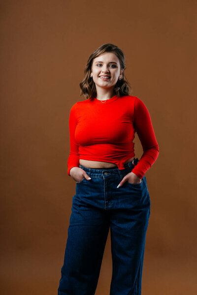 A fashionable young woman in a bold red top and wide-leg denim jeans strikes a dynamic and artistic pose against a warm brown background. High quality photo