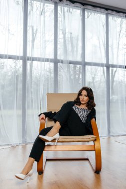 Zarif siyah takım elbiseli şık bir kadın. Taçlı ve beyaz topuklu ayakkabı giyiyor. Modern bir iç mekanda büyük pencereler ve yumuşak doğal ışık ile poz veriyor. Yüksek kalite fotoğraf