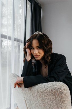 Zarif siyah takım elbiseli şık bir kadın. Taçlı ve beyaz topuklu ayakkabı giyiyor. Modern bir iç mekanda büyük pencereler ve yumuşak doğal ışık ile poz veriyor. Yüksek kalite fotoğraf