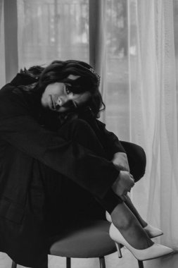 Zarif siyah takım elbiseli şık bir kadın. Taçlı ve beyaz topuklu ayakkabı giyiyor. Modern bir iç mekanda büyük pencereler ve yumuşak doğal ışık ile poz veriyor. Yüksek kalite fotoğraf