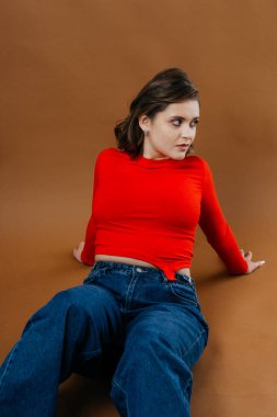 Kalın kırmızı bluzlu ve geniş paçalı kot pantolonlu şık bir genç kadın, dinamik ve sanatsal bir poz veriyor. Yüksek kalite fotoğraf