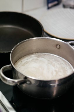 Mutfak malzemelerinin ve çatalların bulanık arkaplanına sahip, paslanmaz çelik bir tencerede yemek pişirirken çekilmiş yakın plan bir fotoğraf. Sıcak bir ev yemeği anı yakalıyor. Yüksek kalite fotoğraf