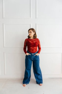 Kırmızı dantelli üst ve geniş bel pantolonlu şık genç bir kadın beyaz panelli bir duvara karşı güven içinde poz veriyor. Yüksek kalite fotoğraf