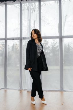 Zarif siyah takım elbiseli şık bir kadın. Taçlı ve beyaz topuklu ayakkabı giyiyor. Modern bir iç mekanda büyük pencereler ve yumuşak doğal ışık ile poz veriyor. Yüksek kalite fotoğraf