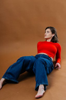 Kalın kırmızı bluzlu ve geniş paçalı kot pantolonlu şık bir genç kadın, dinamik ve sanatsal bir poz veriyor. Yüksek kalite fotoğraf