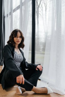 Zarif siyah takım elbiseli şık bir kadın. Taçlı ve beyaz topuklu ayakkabı giyiyor. Modern bir iç mekanda büyük pencereler ve yumuşak doğal ışık ile poz veriyor. Yüksek kalite fotoğraf