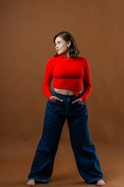 A fashionable young woman in a bold red top and wide-leg denim jeans strikes a dynamic and artistic pose against a warm brown background. High quality photo
