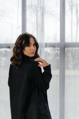 Zarif siyah takım elbiseli şık bir kadın. Taçlı ve beyaz topuklu ayakkabı giyiyor. Modern bir iç mekanda büyük pencereler ve yumuşak doğal ışık ile poz veriyor. Yüksek kalite fotoğraf