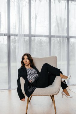 Zarif siyah takım elbiseli şık bir kadın. Taçlı ve beyaz topuklu ayakkabı giyiyor. Modern bir iç mekanda büyük pencereler ve yumuşak doğal ışık ile poz veriyor. Yüksek kalite fotoğraf