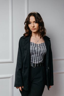 Siyah ceket ve taç giyen kendine güvenen genç bir kadın zarif beyaz bir duvara karşı çapraz kollarla duruyor, modern sofistike ve görkemli cazibeyi harmanlıyor. Yüksek kalite fotoğraf