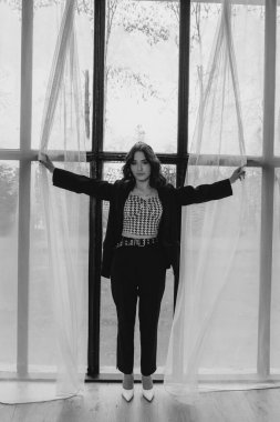 Zarif siyah takım elbiseli şık bir kadın. Taçlı ve beyaz topuklu ayakkabı giyiyor. Modern bir iç mekanda büyük pencereler ve yumuşak doğal ışık ile poz veriyor. Yüksek kalite fotoğraf