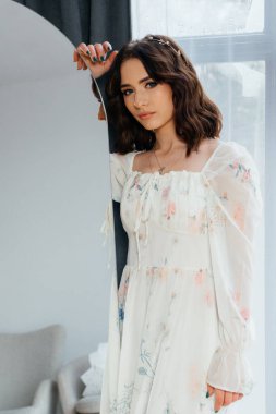 Geniş bir pencerenin yanında, ahşap bir zeminde, dalgalı çiçekli bir elbise giymiş genç bir kadın. Yüksek kalite fotoğraf