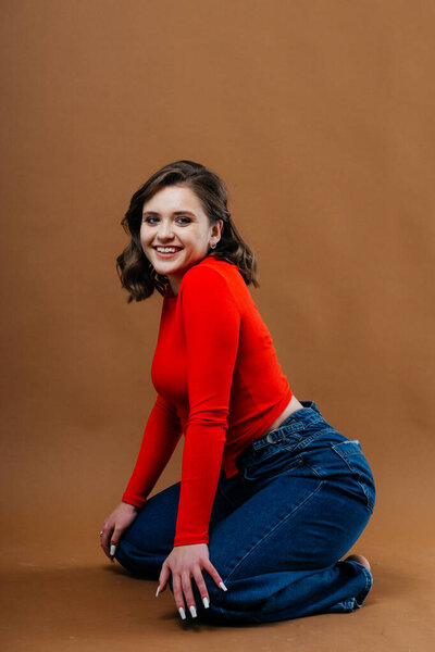 A fashionable young woman in a bold red top and wide-leg denim jeans strikes a dynamic and artistic pose against a warm brown background. High quality photo