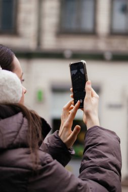 Kışlık ceketli ve kulaklıklı genç bir kadın soğuk bir günde akıllı telefonuyla şehir parkında bir anı yakalıyor. Yüksek kalite fotoğraf