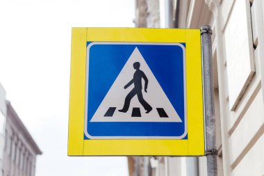 Şehir ortamında bir direğe monte edilmiş mavi ve sarı dizaynlı yaya geçidi tabelası yol güvenliğini ve trafik kurallarını sağlar. Yüksek kalite fotoğraf