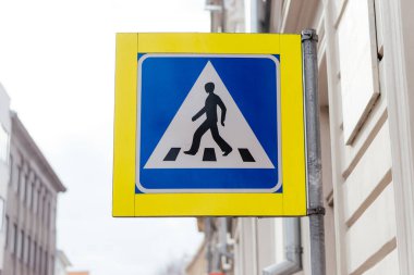 Şehir ortamında bir direğe monte edilmiş mavi ve sarı dizaynlı yaya geçidi tabelası yol güvenliğini ve trafik kurallarını sağlar. Yüksek kalite fotoğraf