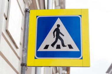 Şehir ortamında bir direğe monte edilmiş mavi ve sarı dizaynlı yaya geçidi tabelası yol güvenliğini ve trafik kurallarını sağlar. Yüksek kalite fotoğraf