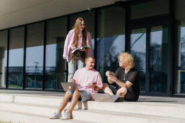 Güneşli bir günde bir grup öğrenci modern bir üniversite binasının önünde dizüstü bilgisayar çalışıyor ve kullanıyor. Yüksek kalite fotoğraf