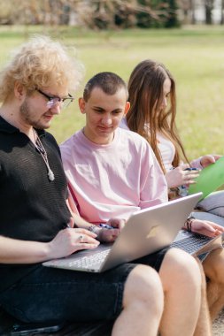 Güneşli bir günde, üç neşeli öğrenci, parkta bir bankta otururken dizüstü bilgisayar ve not defteri kullanarak birlikte çalışır ve birlikte çalışırlar. Yüksek kalite fotoğraf
