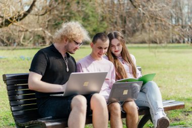 Güneşli bir günde, üç neşeli öğrenci, parkta bir bankta otururken dizüstü bilgisayar ve not defteri kullanarak birlikte çalışır ve birlikte çalışırlar. Yüksek kalite fotoğraf