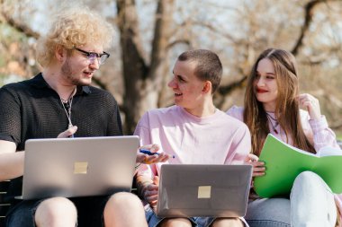 Güneşli bir günde, üç neşeli öğrenci, parkta bir bankta otururken dizüstü bilgisayar ve not defteri kullanarak birlikte çalışır ve birlikte çalışırlar. Yüksek kalite fotoğraf