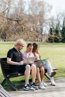 Güneşli bir günde, üç neşeli öğrenci, parkta bir bankta otururken dizüstü bilgisayar ve not defteri kullanarak birlikte çalışır ve birlikte çalışırlar. Yüksek kalite fotoğraf