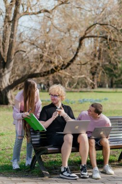 Güneşli bir günde, üç neşeli öğrenci, parkta bir bankta otururken dizüstü bilgisayar ve not defteri kullanarak birlikte çalışır ve birlikte çalışırlar. Yüksek kalite fotoğraf