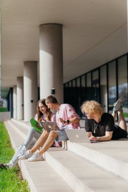 Güneşli bir günde bir grup öğrenci modern bir üniversite binasının önünde dizüstü bilgisayar çalışıyor ve kullanıyor. Yüksek kalite fotoğraf