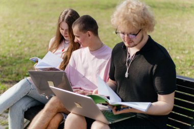 Güneşli bir günde, üç neşeli öğrenci, parkta bir bankta otururken dizüstü bilgisayar ve not defteri kullanarak birlikte çalışır ve birlikte çalışırlar. Yüksek kalite fotoğraf