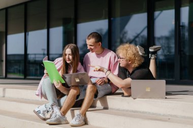 Güneşli bir günde bir grup öğrenci modern bir üniversite binasının önünde dizüstü bilgisayar çalışıyor ve kullanıyor. Yüksek kalite fotoğraf