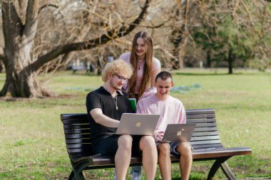 Güneşli bir günde, üç neşeli öğrenci, parkta bir bankta otururken dizüstü bilgisayar ve not defteri kullanarak birlikte çalışır ve birlikte çalışırlar. Yüksek kalite fotoğraf