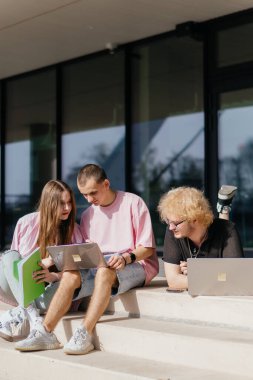 Güneşli bir günde bir grup öğrenci modern bir üniversite binasının önünde dizüstü bilgisayar çalışıyor ve kullanıyor. Yüksek kalite fotoğraf