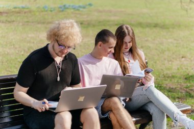 Güneşli bir günde, üç neşeli öğrenci, parkta bir bankta otururken dizüstü bilgisayar ve not defteri kullanarak birlikte çalışır ve birlikte çalışırlar. Yüksek kalite fotoğraf