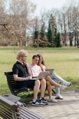Güneşli bir günde, üç neşeli öğrenci, parkta bir bankta otururken dizüstü bilgisayar ve not defteri kullanarak birlikte çalışır ve birlikte çalışırlar. Yüksek kalite fotoğraf