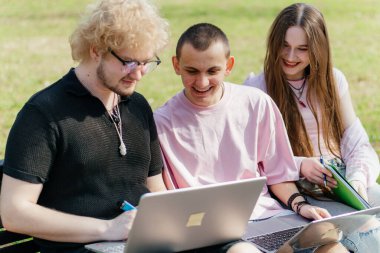 Güneşli bir günde, üç neşeli öğrenci, parkta bir bankta otururken dizüstü bilgisayar ve not defteri kullanarak birlikte çalışır ve birlikte çalışırlar. Yüksek kalite fotoğraf