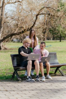 Güneşli bir günde, üç neşeli öğrenci, parkta bir bankta otururken dizüstü bilgisayar ve not defteri kullanarak birlikte çalışır ve birlikte çalışırlar. Yüksek kalite fotoğraf
