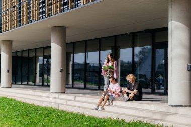 Güneşli bir günde bir grup öğrenci modern bir üniversite binasının önünde dizüstü bilgisayar çalışıyor ve kullanıyor. Yüksek kalite fotoğraf