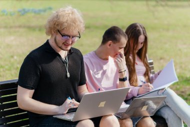 Güneşli bir günde, üç neşeli öğrenci, parkta bir bankta otururken dizüstü bilgisayar ve not defteri kullanarak birlikte çalışır ve birlikte çalışırlar. Yüksek kalite fotoğraf