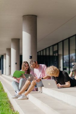 Güneşli bir günde bir grup öğrenci modern bir üniversite binasının önünde dizüstü bilgisayar çalışıyor ve kullanıyor. Yüksek kalite fotoğraf