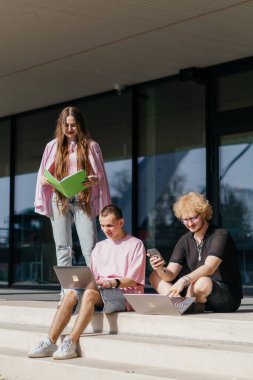 Güneşli bir günde bir grup öğrenci modern bir üniversite binasının önünde dizüstü bilgisayar çalışıyor ve kullanıyor. Yüksek kalite fotoğraf